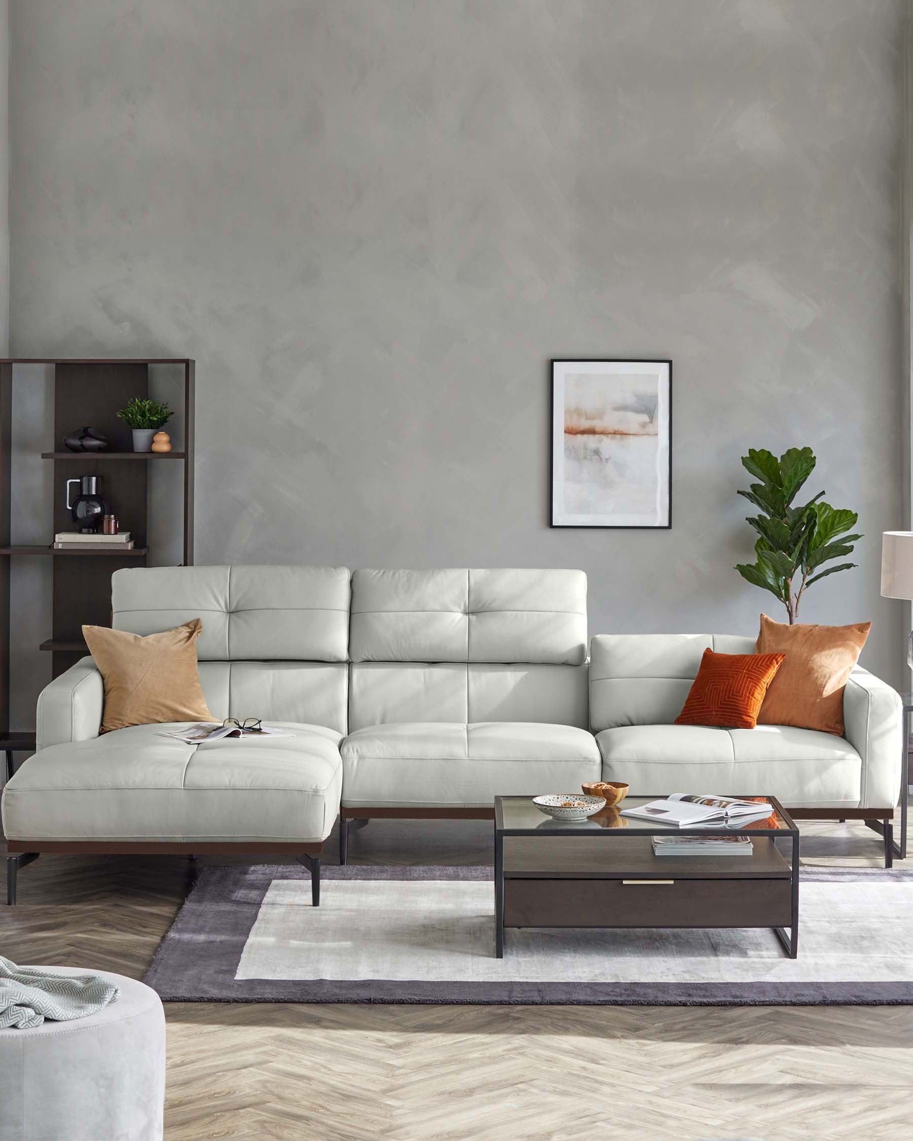 Modern living room setup featuring a light grey L-shaped sectional sofa with plush back cushions, accented with tan and orange throw pillows. In front of the sofa is a rectangular dark wood coffee table with a lower shelf and glass top, holding decorative items and books. To the left, a dark wooden shelving unit displaying minimalistic decor complements the furniture ensemble. A soft grey area rug underfoot adds warmth to the arrangement.