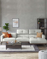 Modern living room setup featuring a light grey L-shaped sectional sofa with clean lines and plush cushions, accented with burnt orange throw pillows. In front of the sofa is a rectangular dark wood coffee table with a lower shelf, displaying books and decorative items. To the side, there's a dark wood shelving unit with neatly arranged accessories. The furniture rests on a striped grey and white area rug, complementing the room's neutral colour palette.