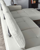 Modern light grey sectional sofa with a tufted backrest and cushioned seats, featuring clean lines and chrome-plated sleek legs, partially visible in a contemporary living room setting with a grey wall and wood flooring. A dark wooden shelf with decorative items is in the background.