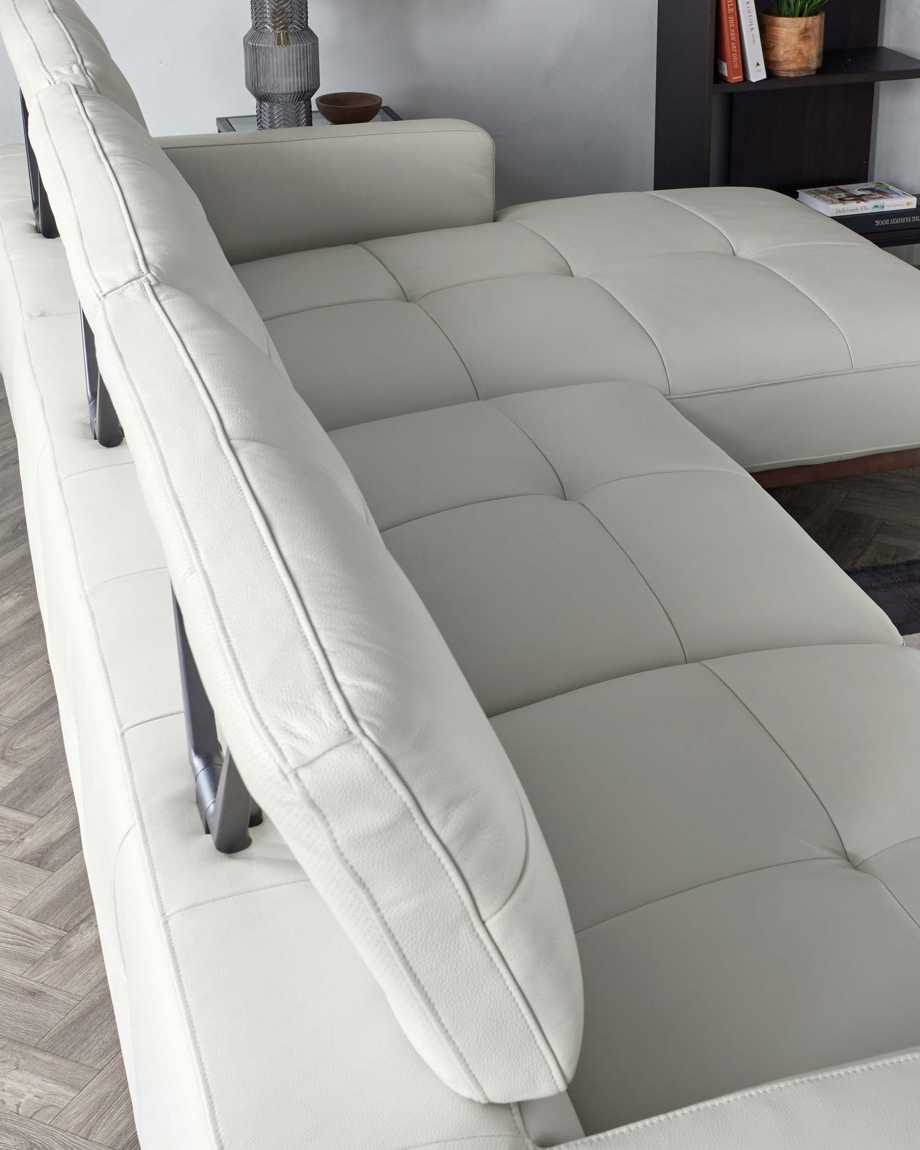 A modern light-grey sectional sofa with plush cushions and sleek lines, featuring a tufted backrest and seat design with visible stitching details. It rests on slim, elevated metal legs, adding to its contemporary aesthetic. The scene includes a partial view of a bookshelf and decorative items in the background, complementing the minimalist style of the room.