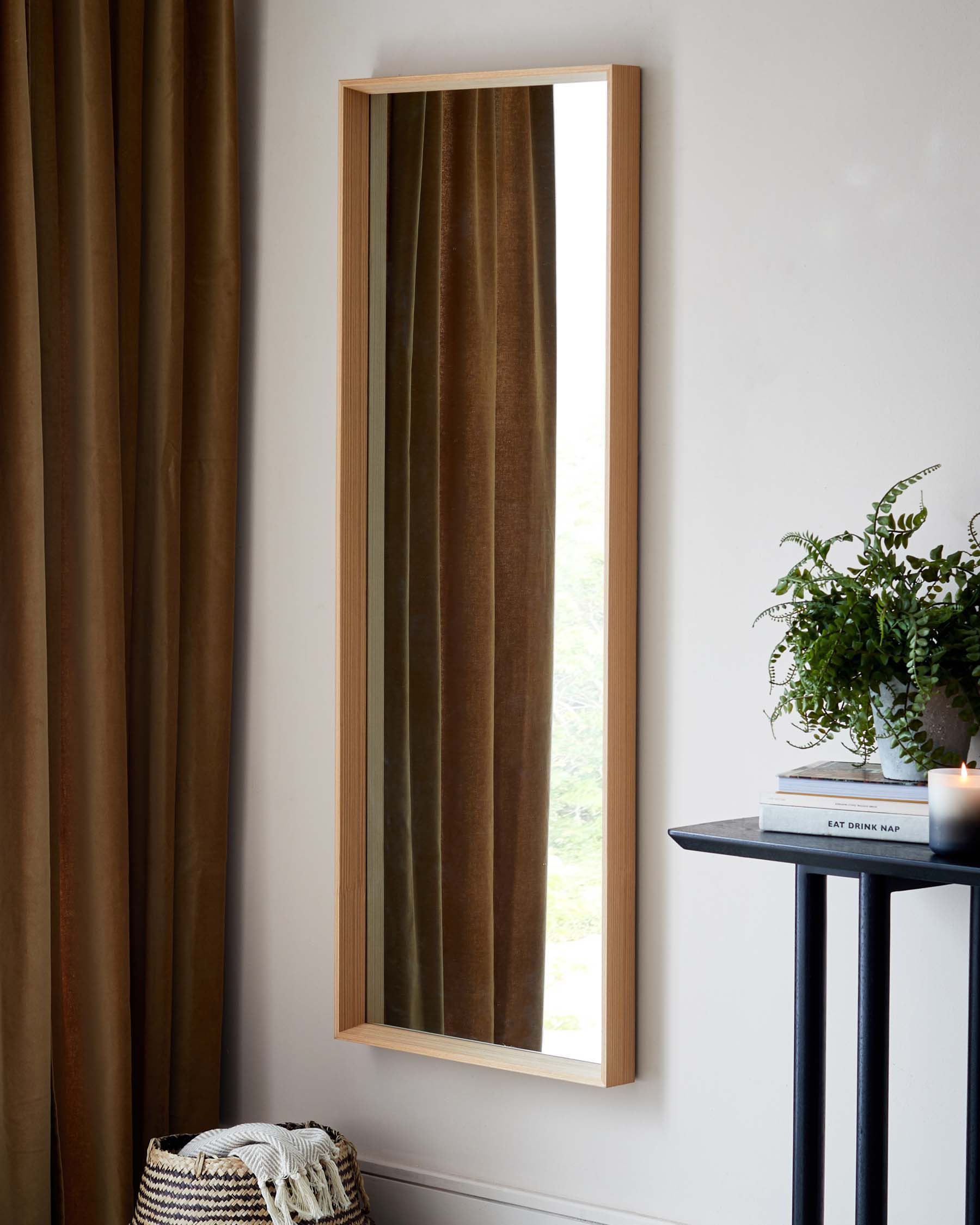 A sleek, modern tall mirror with a light natural wood frame, mounted vertically on a white wall, alongside a minimalist black side table with a round top and slender metal legs. The table displays a potted fern, a stack of books, and a small lit candle, creating a cosy and contemporary atmosphere.