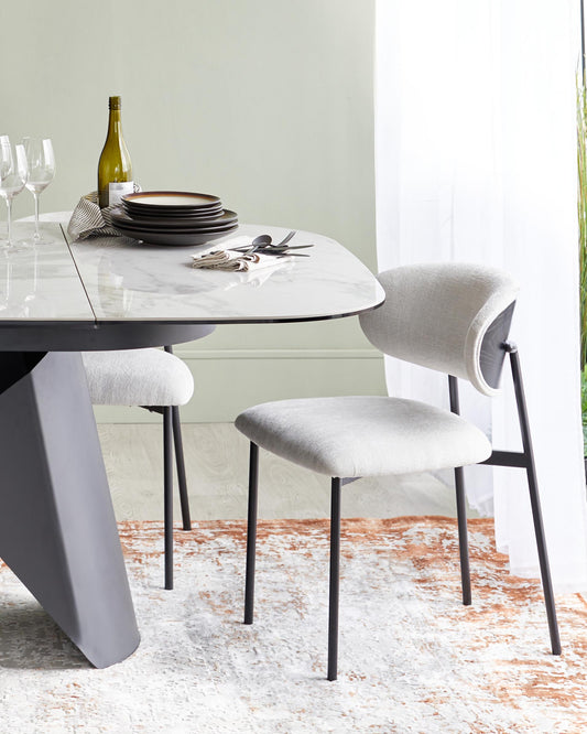 A modern dining table with a marble top and sleek black base, paired with light grey upholstered chairs on a textured rug.