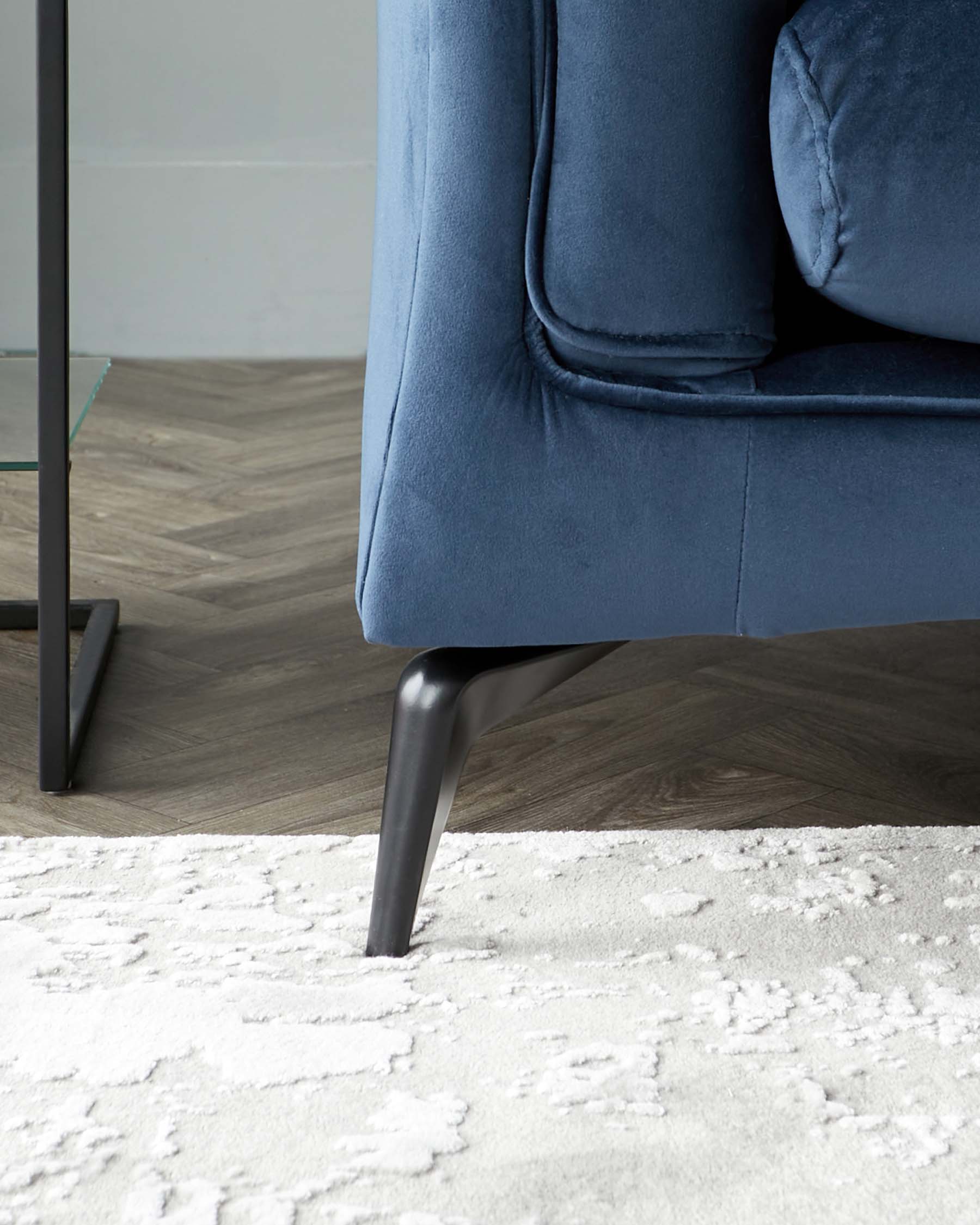 A plush blue velvet sofa with sleek black legs, placed on a textured white rug over a stylish herringbone floor.