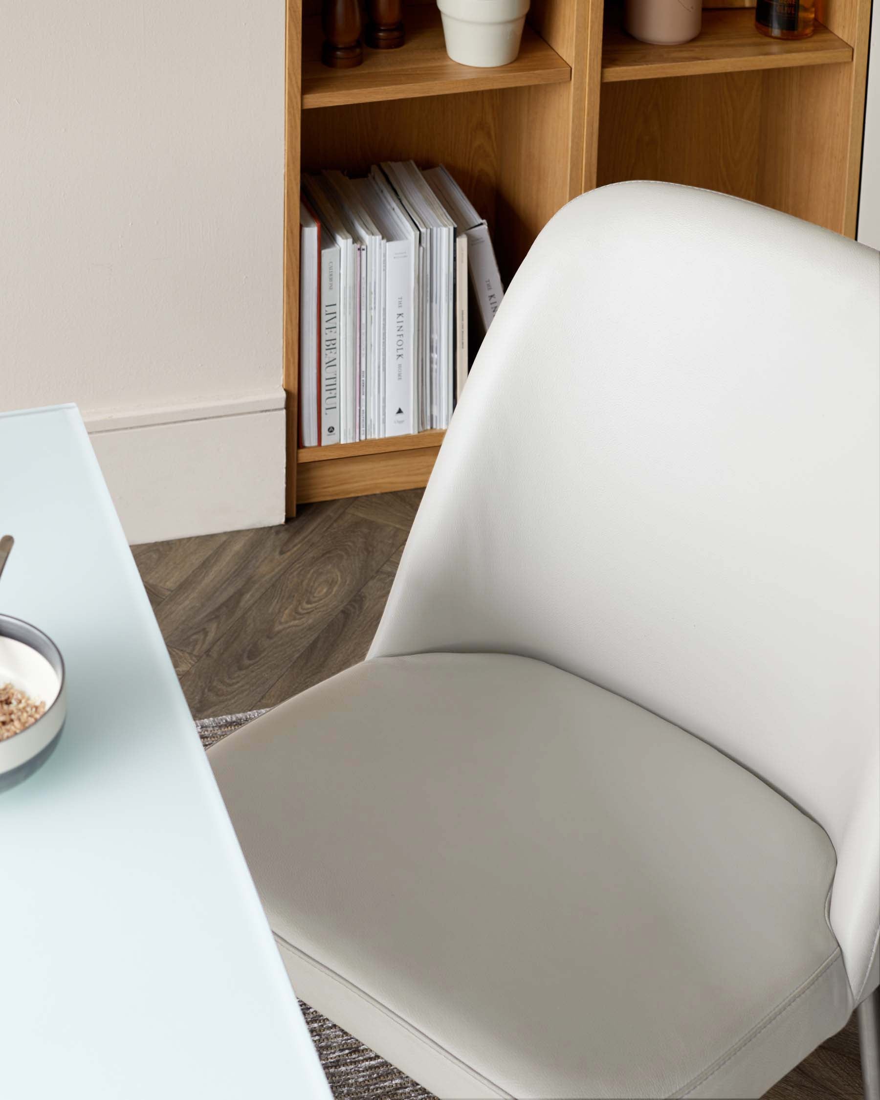 A sleek white chair beside a glass table, featuring a minimalist design and a natural wood shelving unit in the background.