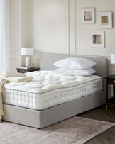 Elegant bedroom setup featuring a plush mattress, light gray upholstered bed frame, nightstand, lamp, and decorative wall art.