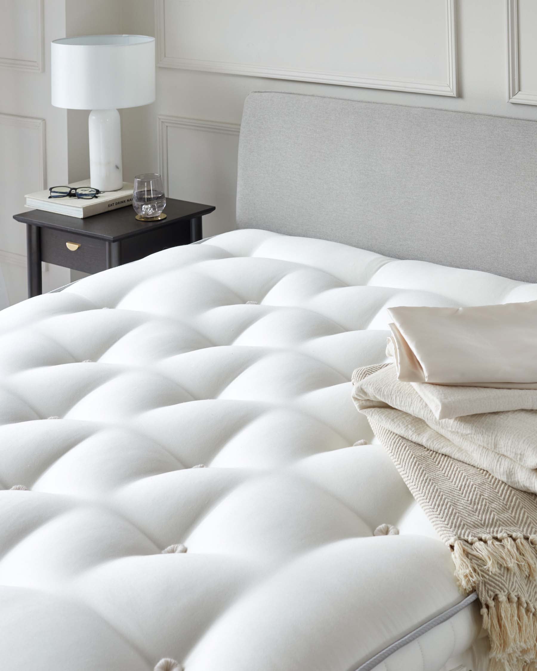 Elegant bedroom setup featuring a plush white mattress, soft linens, a chic gray headboard, and a stylish black nightstand with a lamp.