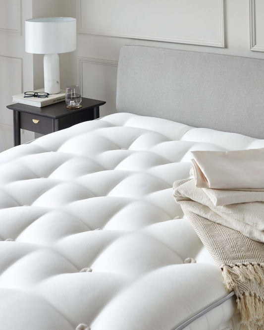 A plush, tufted white mattress with a light gray headboard, accompanied by a dark nightstand, lamp, and folded blankets.