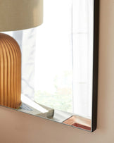 Part of a beige fabric lampshade over a ribbed amber base, reflecting in a mirror with a black frame next to a sheer white curtain by a sunlit window.