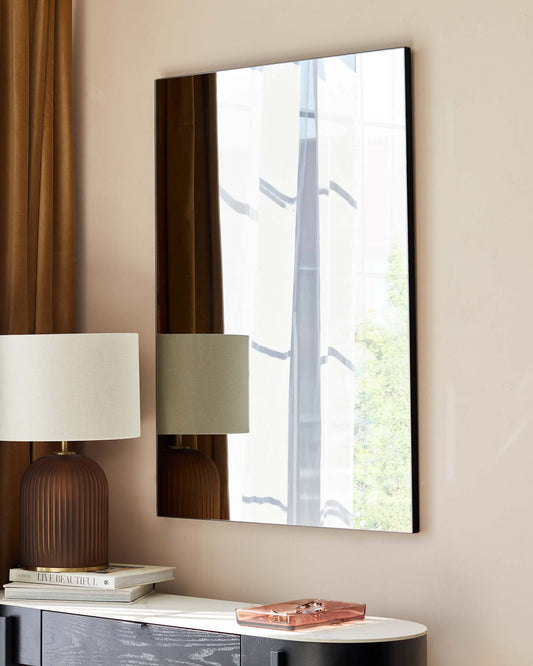 A contemporary room featuring a sleek, white and black console table with a decorative black drawer, flanked by two elegant ribbed, dark brown table lamps with cream lampshades. A neat stack of hardcover books and a stylish pink decorative box rest atop the console table, enhancing the modern aesthetic of the space. A large wall mirror above reflects the room's natural light.