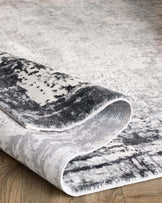 A plush modern area rug with a rolled corner, featuring a grayscale abstract pattern, displayed on a wooden floor.