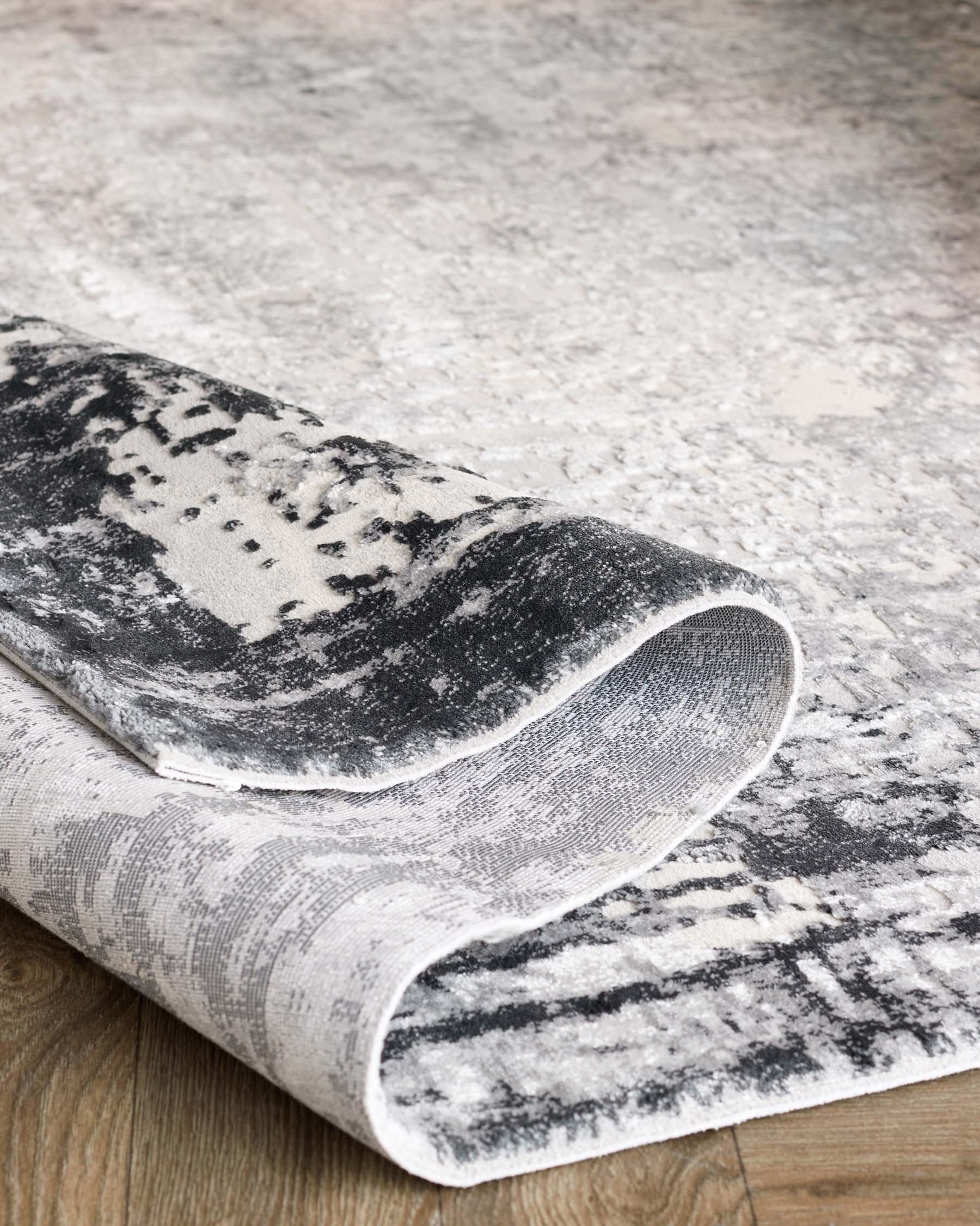 Elegant modern area rug featuring a distressed grayscale pattern with a rolled corner, showcasing the rug's texture and thickness, laid out on a wooden floor.