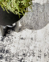 Contemporary round side table with a black finish, supported by a unique Y-shaped base. The table is partially obscured by a hanging potted plant with green foliage, and is positioned on a textured area rug with an abstract black and white pattern.