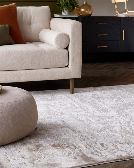 Beige modern sofa with cylindrical bolster pillow, matching fabric ottoman, and black wooden credenza with sleek brass handles on a plush area rug.