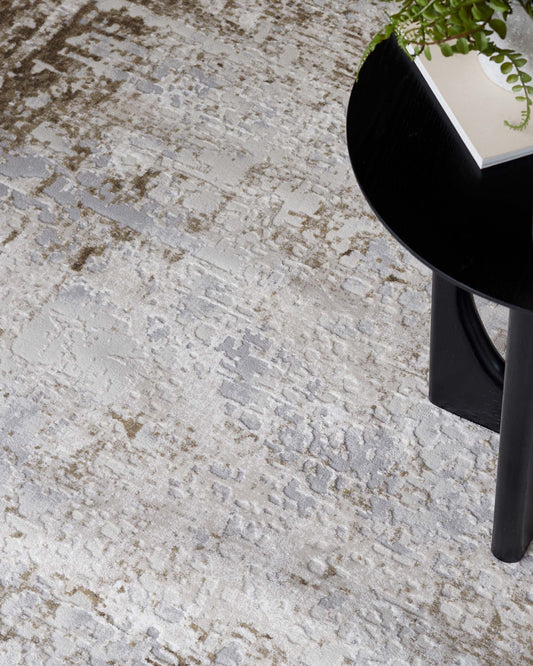 A modern black circular side table with a sleek finish and a minimalist design, featuring a thin vertical base and topped with a book and a potted plant. The table is set against a textured cream and grey rug with an abstract pattern.