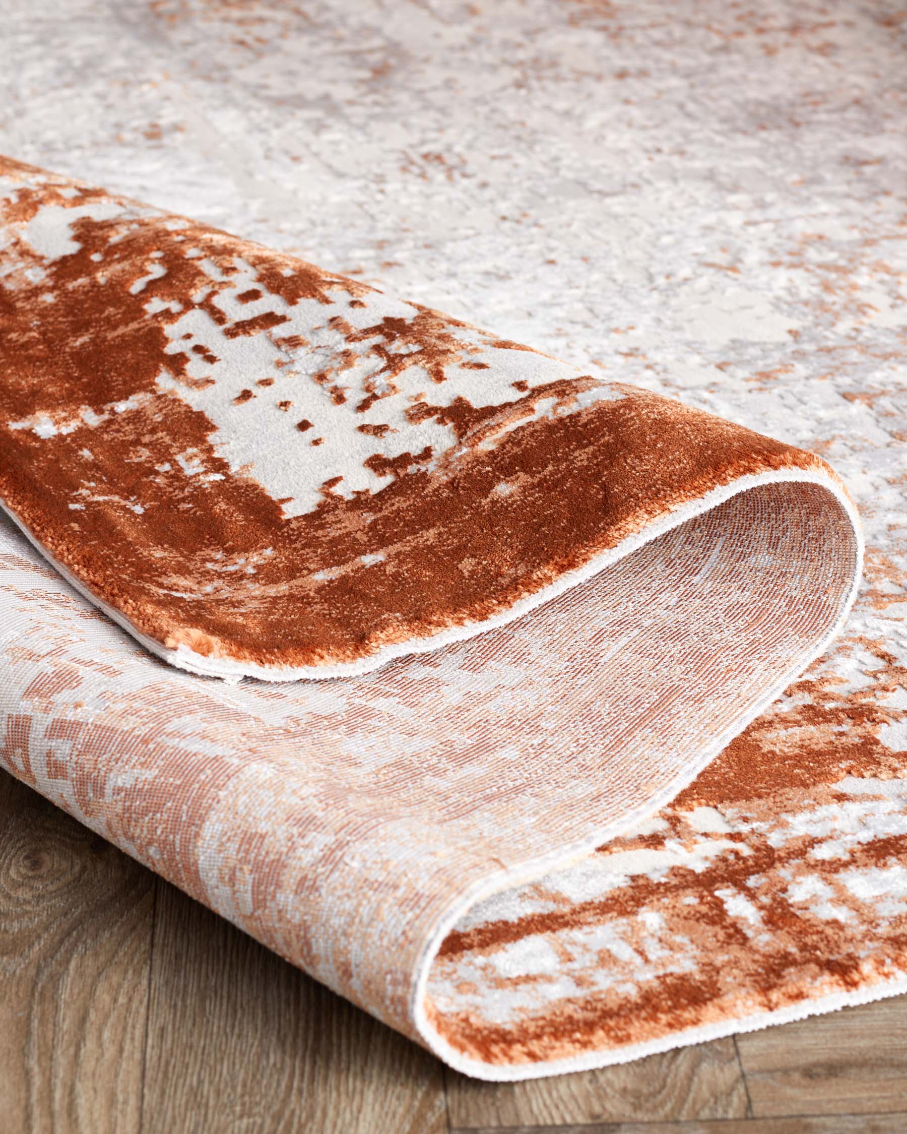 Elegant area rug with a white and burnt orange distressed pattern, partially rolled up to showcase its texture, set against a wooden floor background.