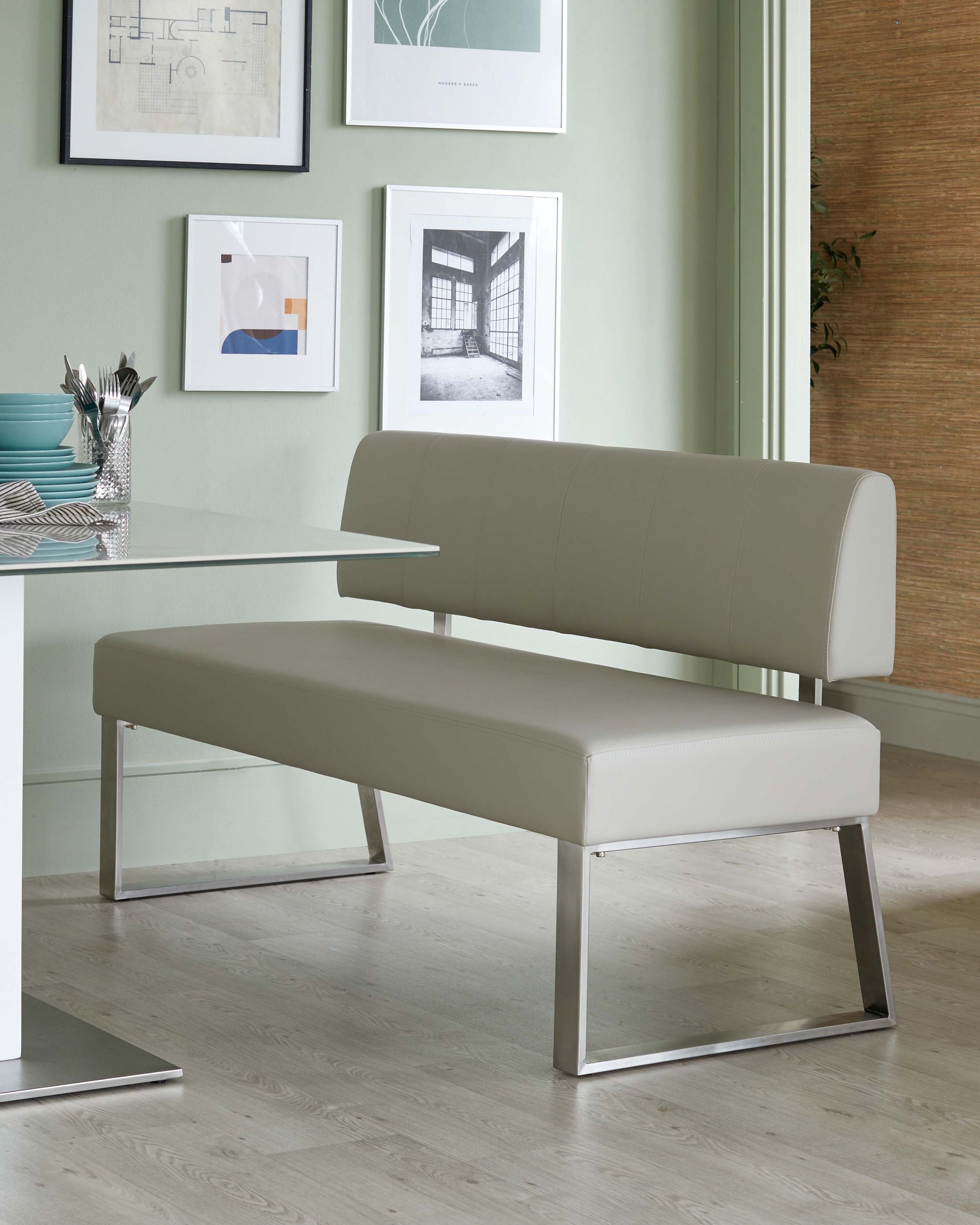 Modern minimalist bench with a tufted, light grey upholstered seat and backrest, featuring sleek, metallic chrome legs in a geometric design. The bench is paired with a simple white rectangular table with a similar chrome leg finish. The room has a contemporary aesthetic with wall art and light hardwood flooring.