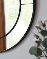 The image does not contain any furniture. It displays a large round wall mirror with a black frame, partially obscured on the right side by out-of-focus white flowers and green leaves of an indoor plant. The mirror reflects a view of trees and greenery outside a window.