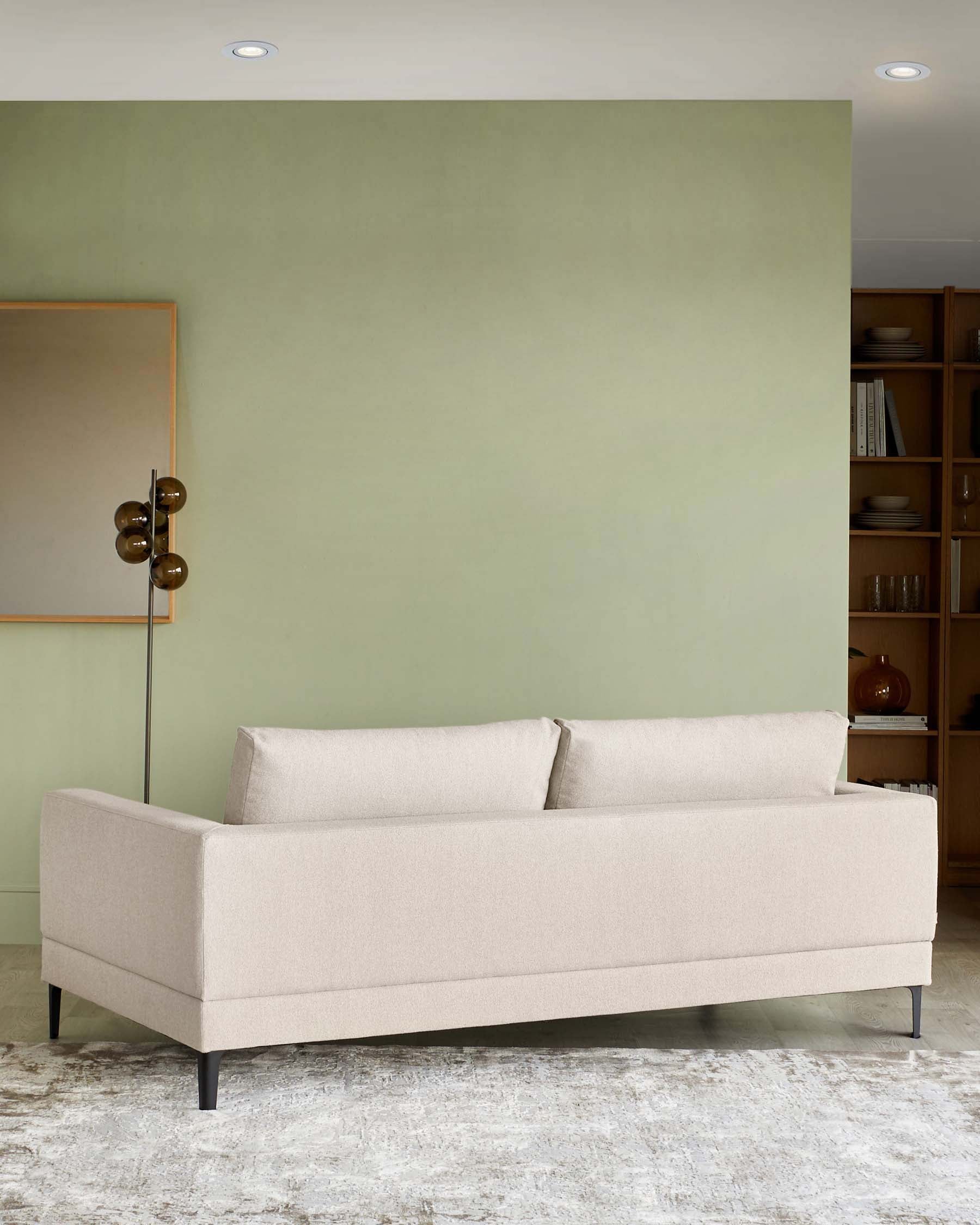 A contemporary beige three-seater sofa with a clean, simple design, featuring slim arms and black metal legs, set against a neutral-toned rug on a hardwood floor. A tall, mid-century-inspired floor lamp with a bronze finish and spherical shades can be seen to the left, next to a large, framed mirror with a metallic border, and a wooden bookshelf filled with various items occupies the right side of the image.