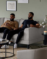 A modern, two-seater light grey fabric sofa with clean lines and dark, low-profile legs. In front of the sofa, there's a round coffee table featuring a glass top and a black metal frame. On the right side of the sofa stands a sleek, dark-toned round side table with a candle on it, harmonizing with the room's contemporary style. The room's decor is completed by a striped area rug in neutral tones that complement the furniture's colour palette.