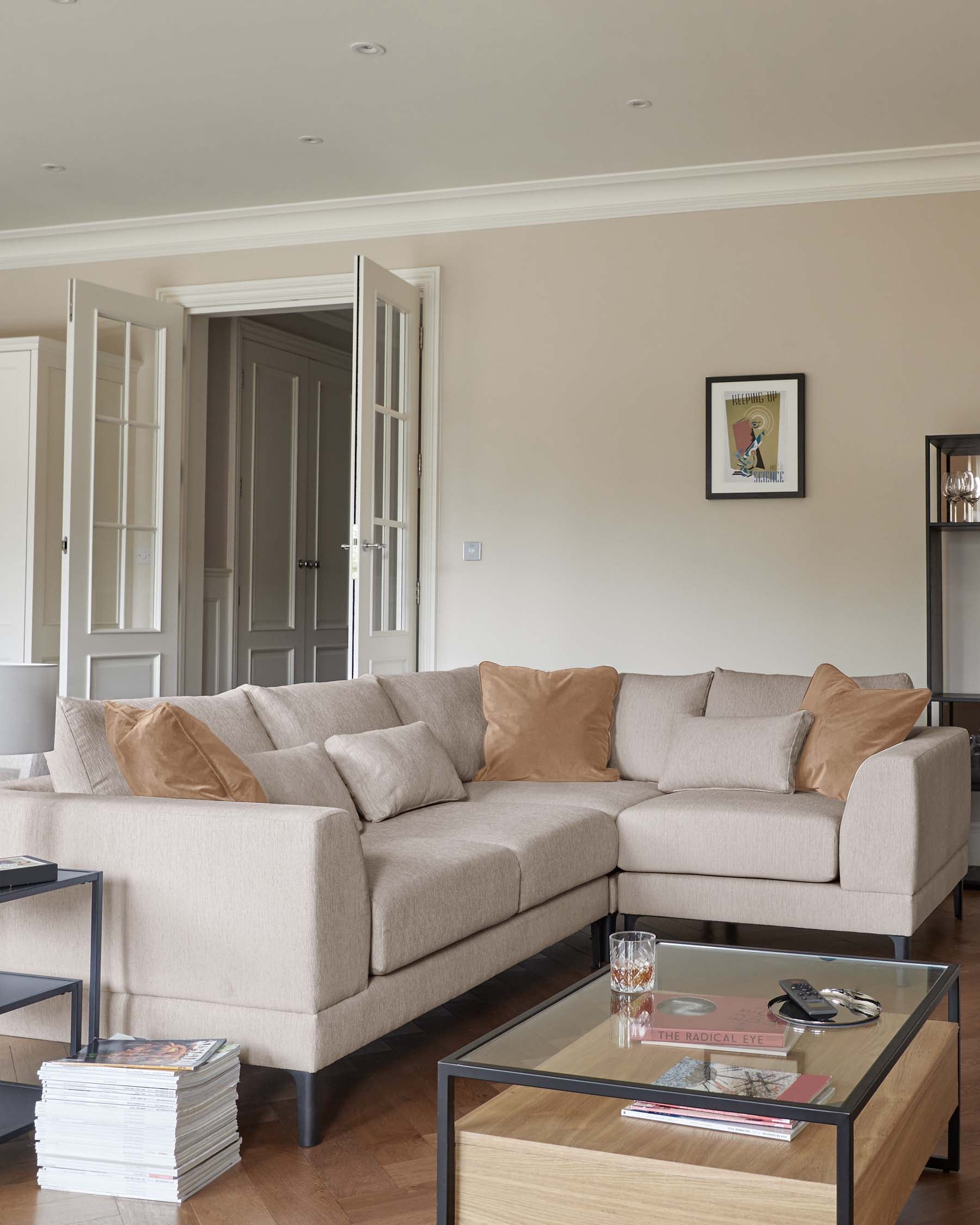 Light beige sectional sofa with cushions, a glass-top coffee table with wooden base, and stacked magazines beside it.