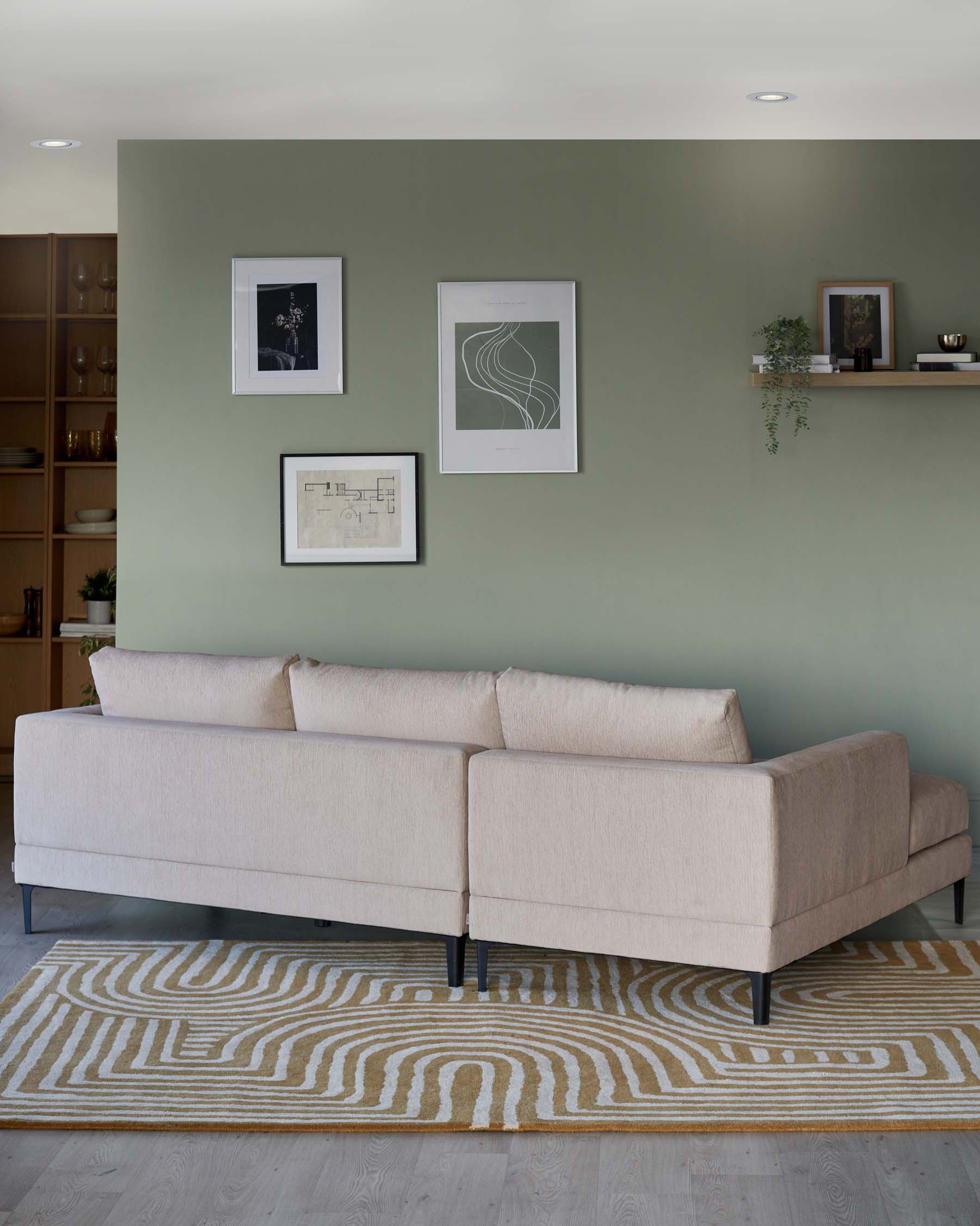 A modern light beige sectional sofa with clean lines and dark slender legs, sitting on a contemporary patterned rug in beige and white tones. Behind the sofa, a minimalist wood shelving unit displays decorative items, all against a pale green wall adorned with framed art.