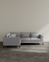 Modern light grey fabric sectional sofa with simple lines, three seat cushions, and two matching rectangular throw pillows, elevated on dark wooden legs, set in a minimalist room with herringbone-patterned wood flooring.