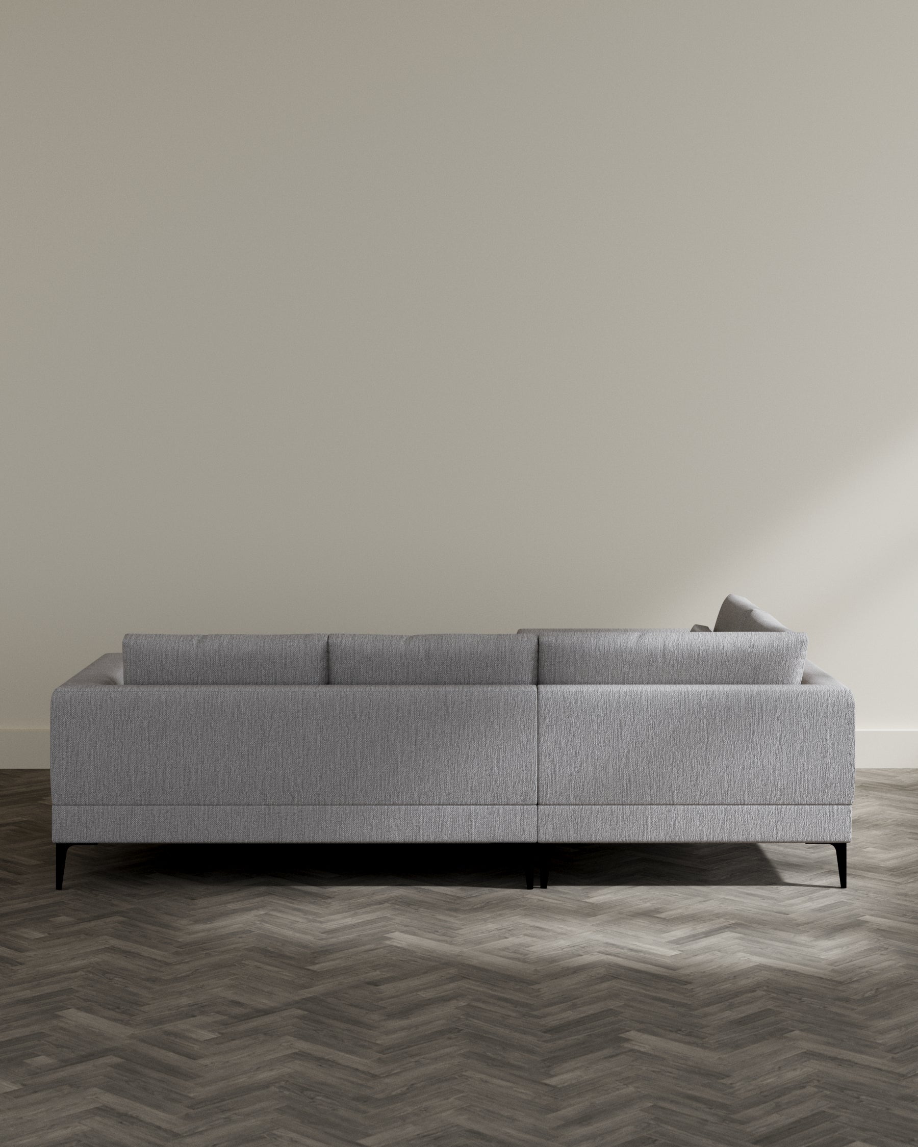 A modern three-seater sofa with a minimalist design, featured in a monochromatic grey fabric upholstery with a subtle texture. The sofa is elevated on low-profile black legs, which provide a sleek contrast to the light upholstery. It has a clean-lined, boxy silhouette with three back cushions and a single long seat cushion that emphasizes comfort without sacrificing style. The sofa is positioned on a herringbone-patterned wood floor, adding warmth to the overall contemporary aesthetic of the piece.