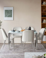 A sleek, modern dining set featuring a glass-top table and five upholstered white chairs with metallic legs, set on a patterned rug.