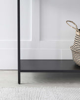 A sleek black shelf on slim legs, paired with a woven basket, set against a light gray rug and minimalistic wall backdrop.