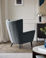 A modern wingback armchair upholstered in dark grey fabric with a curved backrest and angled wooden legs, positioned next to a sideboard cabinet with vertical black ribbing and a round, white marble-topped coffee table in the foreground. The scene is complemented by minimalist decor, including a framed abstract painting, metal spherical table lamp, and a book stack with a candle atop the cabinet.