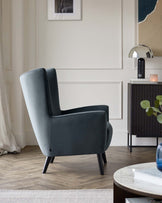 Elegant modern living room with a slate grey wingback chair with tapered dark wooden legs, positioned next to a black cabinet with slatted front design and sleek metal handles. The cabinet is topped with a small silver lamp and a lit candle, emphasizing a contemporary and sophisticated interior style. Further decor includes a round coffee table with books, a framed picture on the wall, light drapery, and a potted plant, creating a relaxed and stylish ambiance.