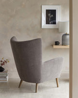 A modern high-back wing chair with a sleek profile, upholstered in textured grey fabric, supported by splayed wooden legs, positioned beside a small wooden shelf displaying a decorative lamp and framed artwork.