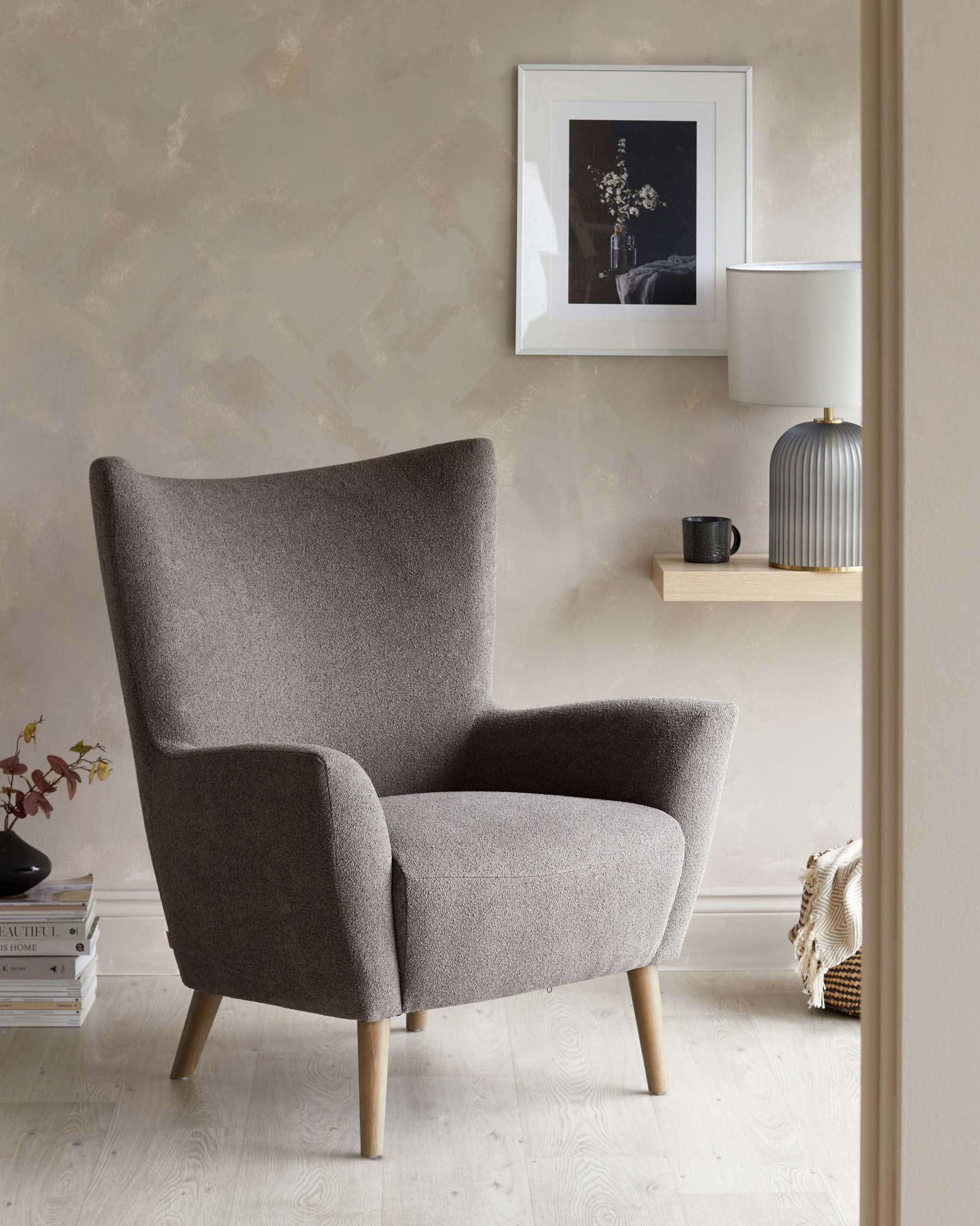 Modern high-back armchair with textured grey upholstery and light wooden legs placed in a cosy corner of a room with neutral-toned walls. A small wooden shelf with a decorative lamp and a framed artwork above it complement the minimalist decor. A stack of books and a woven basket with a throw blanket are nearby, providing a lived-in ambiance.