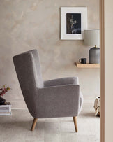 Modern grey fabric upholstered armchair with a high backrest and angled wooden legs, positioned in a cosy room with neutral-toned decor.