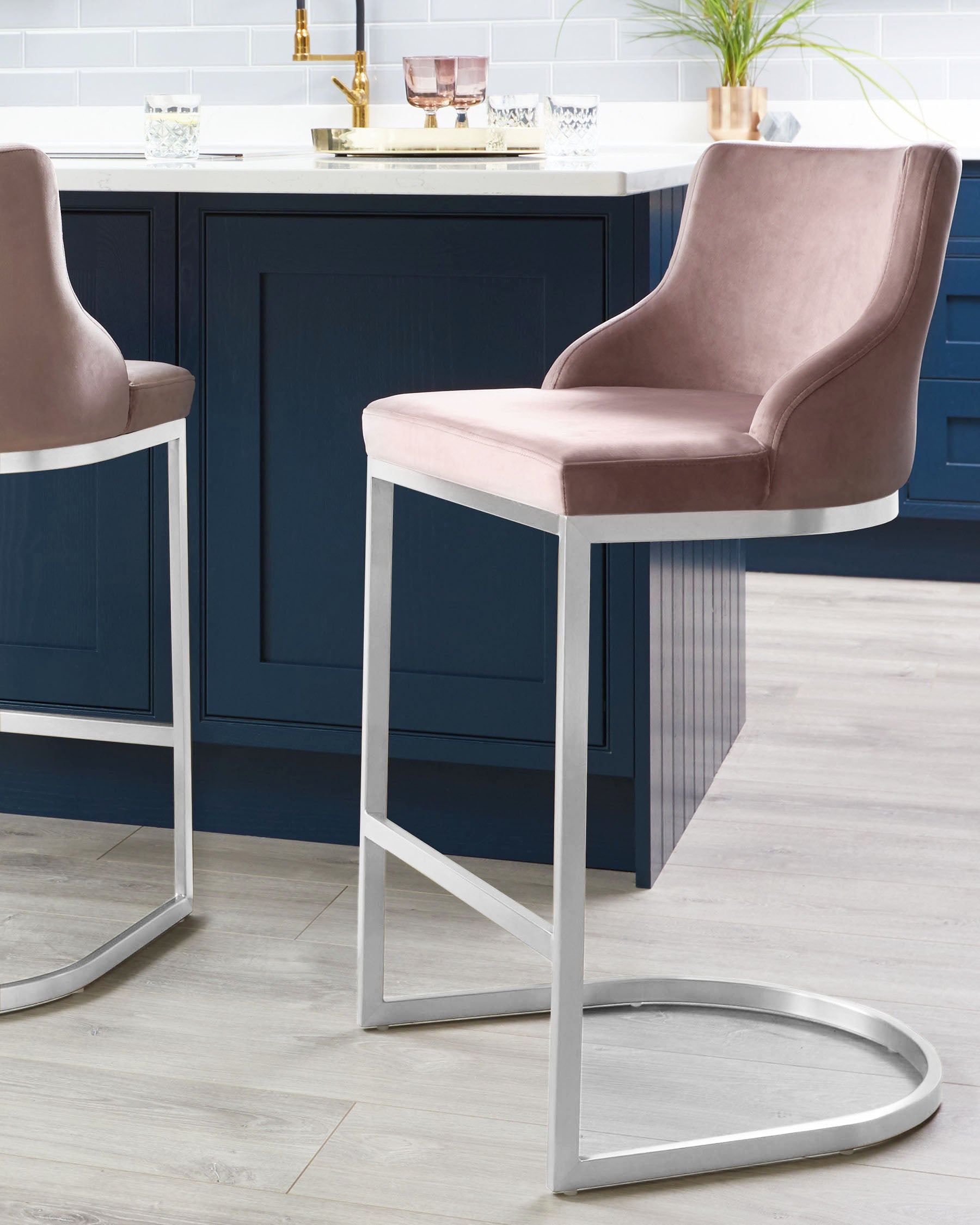 Two elegant velvet counter stools with silver metal frames, positioned near a modern kitchen island with a dark blue finish.