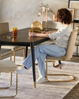 Elegant dining room set featuring a dark wooden table with black legs and gold accents, and a set of beige upholstered chairs with gold metal frames. A plush light rug lies underneath, complementing the warm neutral tones of the room.