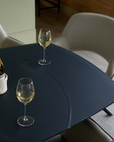 A sleek dark table with a curved edge, accompanied by modern light-colored chairs and wine glasses on top.