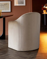 A soft, curved chair in light fabric, positioned next to a modern wooden table, with warm ambient lighting in a stylish setting.