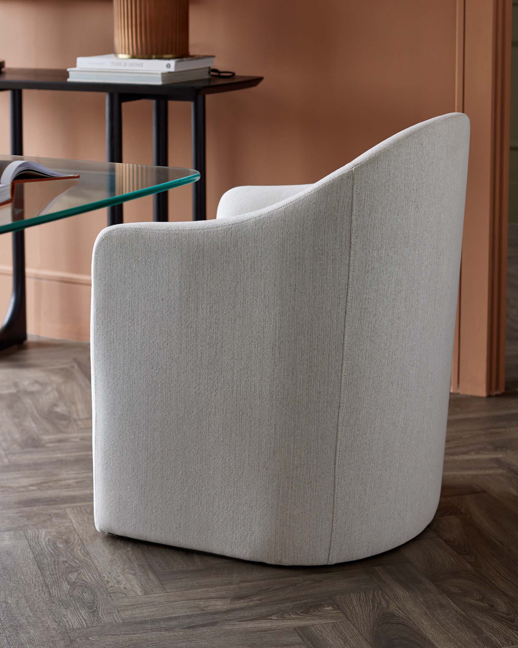 A sleek, modern armchair in light gray fabric, paired with a glass side table and set against a warm, earthy wall.