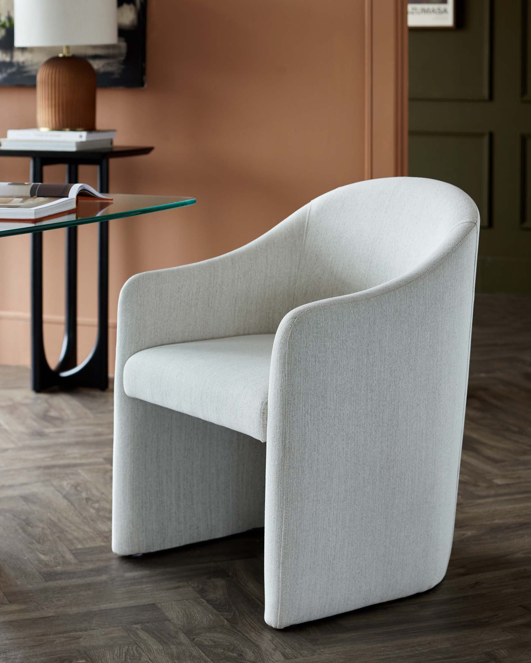 A stylish light gray upholstered chair with rounded edges, paired with a glass top table and a decorative lamp in a modern setting.