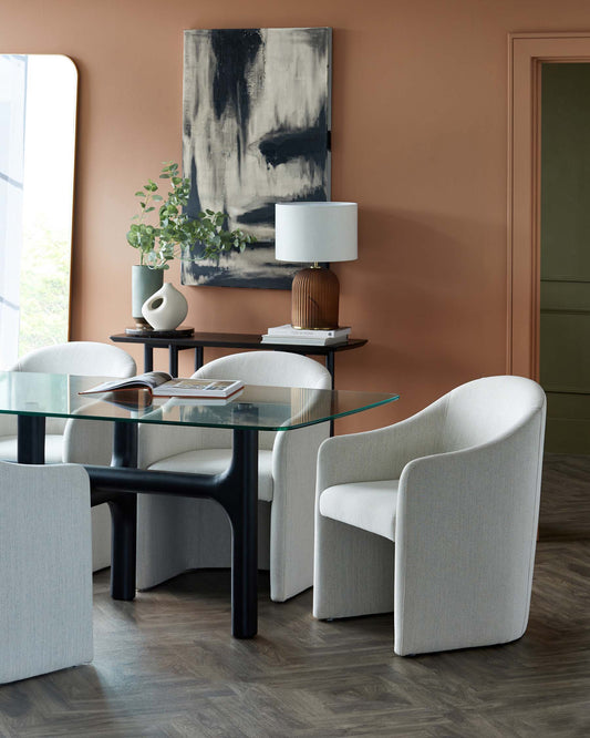 A modern dining setup featuring four rounded white chairs, a glass-top table, and an elegant vase with greenery on a dark console.