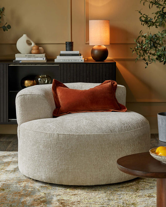 A cozy circular chair with a textured beige fabric and an orange velvet pillow, accompanied by a dark wood side table and lamp.