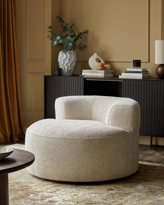 A cozy, round, ivory accent chair, paired with a modern side table and a stylish console displaying decorative items and books.