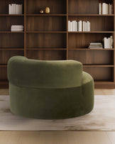 A rounded green velvet chair stands before a wooden bookshelf filled with white books, creating a cozy, modern atmosphere.