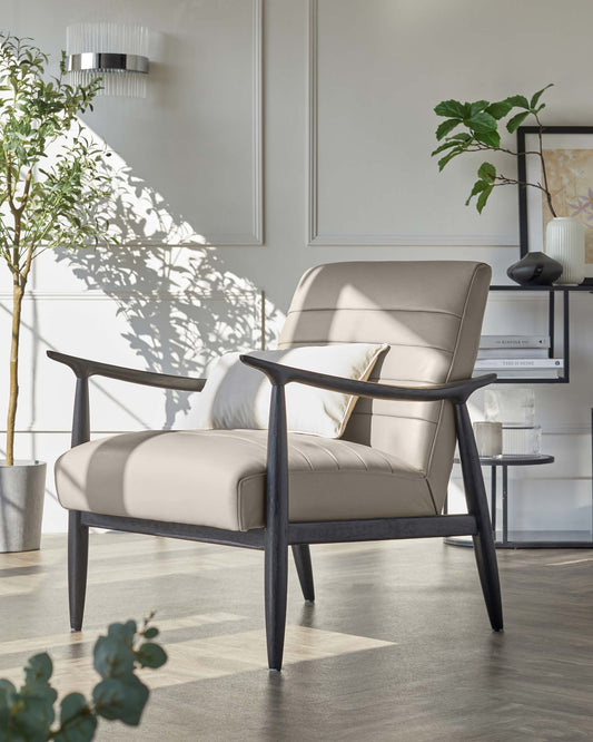 Modern armchair with a beige upholstered seat and back cushion, featuring a solid dark wooden frame with sleek, angular arms. A white decorative pillow rests on the seat. In the background, a minimalistic black-framed bookshelf holds a variety of items including books, a vase, and decorative objects, adjacent to a potted indoor plant. A stylish round wall lamp emits soft lighting, casting a shadow of the plant on the wall.