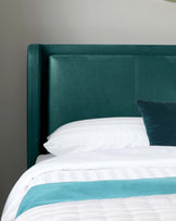 Teal upholstered headboard with a simple, modern design featuring clean lines and white stitching detail, accompanied by a matching teal velvet throw pillow resting on a bed with crisp white linens and a turquoise accent stripe.