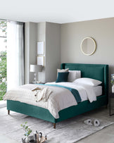 Elegant teal upholstered bed frame with a high headboard and matching footboard, accented by tapered wooden legs. A modern bedside table in black with a sleek lamp and a round, gold-framed wall mirror complement the contemporary bedroom aesthetic.