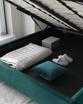 Contemporary style upholstered storage bed in teal with a lifted base showing storage space underneath, complemented by a plush white comforter, teal and grey throw pillows, and a pair of white slippers at the foot of the bed.