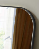 Partial view of a sleek, modern mirror with a curved top and wooden backing, featuring a thin, dark metal frame mounted on a light-coloured wall.