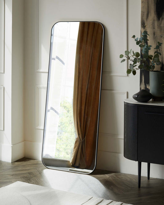 A modern full-length floor mirror with a rounded top and black frame leaning against a white panelled wall. To its right is a minimalist black sideboard with subtle wood grain texture, standing on slender, tapered legs. A potted plant with silvery green foliage is perched atop the sideboard next to an understated black vase.